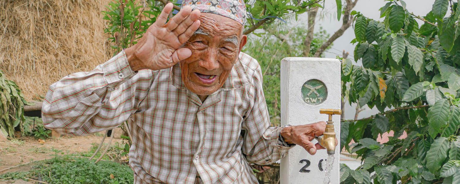 Elderly Gurkha at tap stand saluting and grinning in Nepal